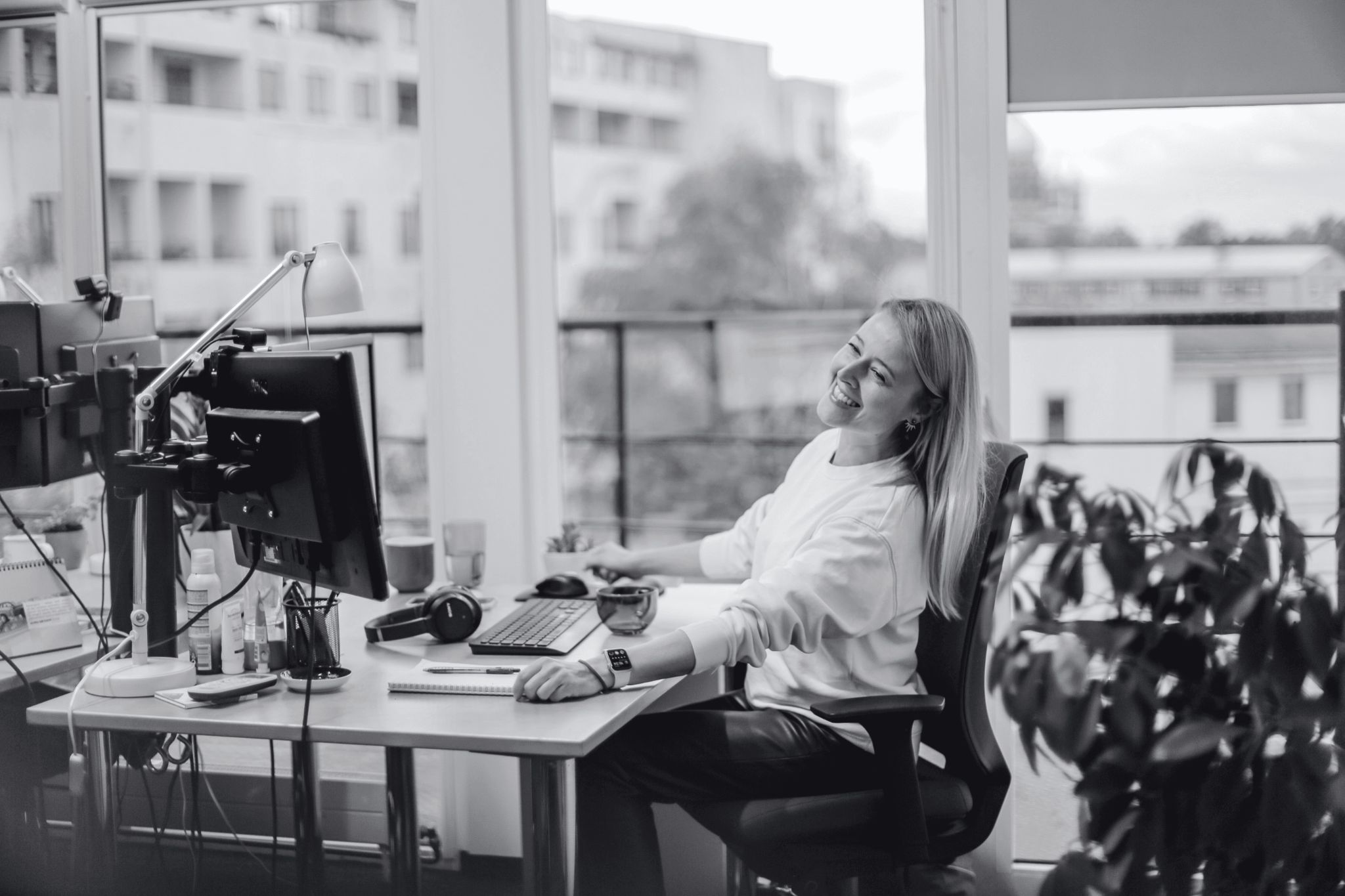 A person sitting at a desk in an office, looking back with a smile, surrounded by colleagues and natural light.