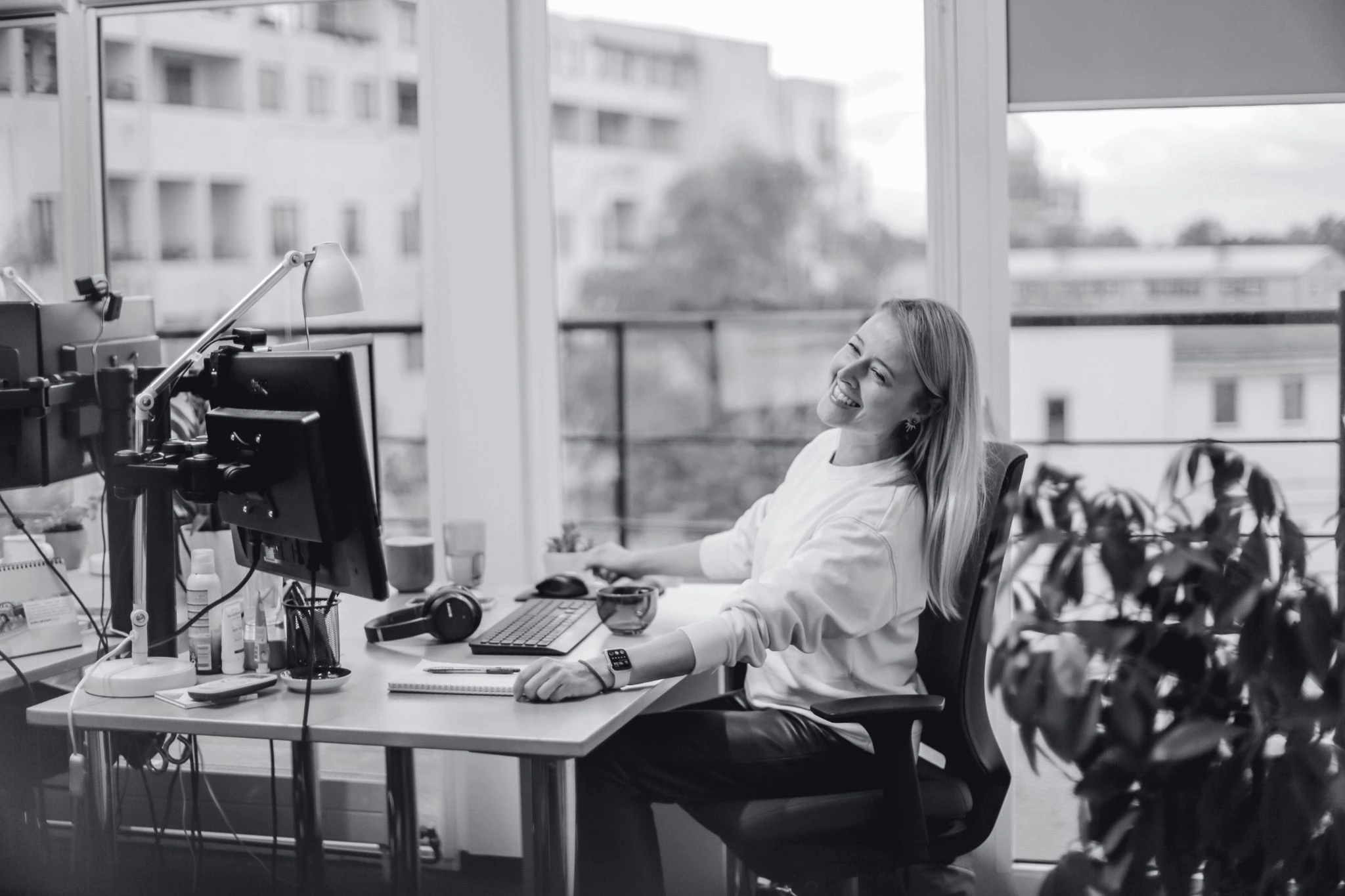 A person sitting at a desk in an office, looking back with a smile, surrounded by colleagues and natural light.