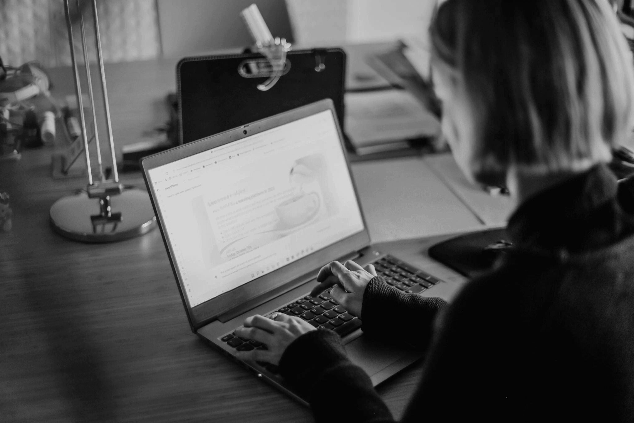 A person working on a laptop at a desk, viewing a presentation about a coffee cup, with a modern lamp and a clipboard in the background, in a monochrome setting.