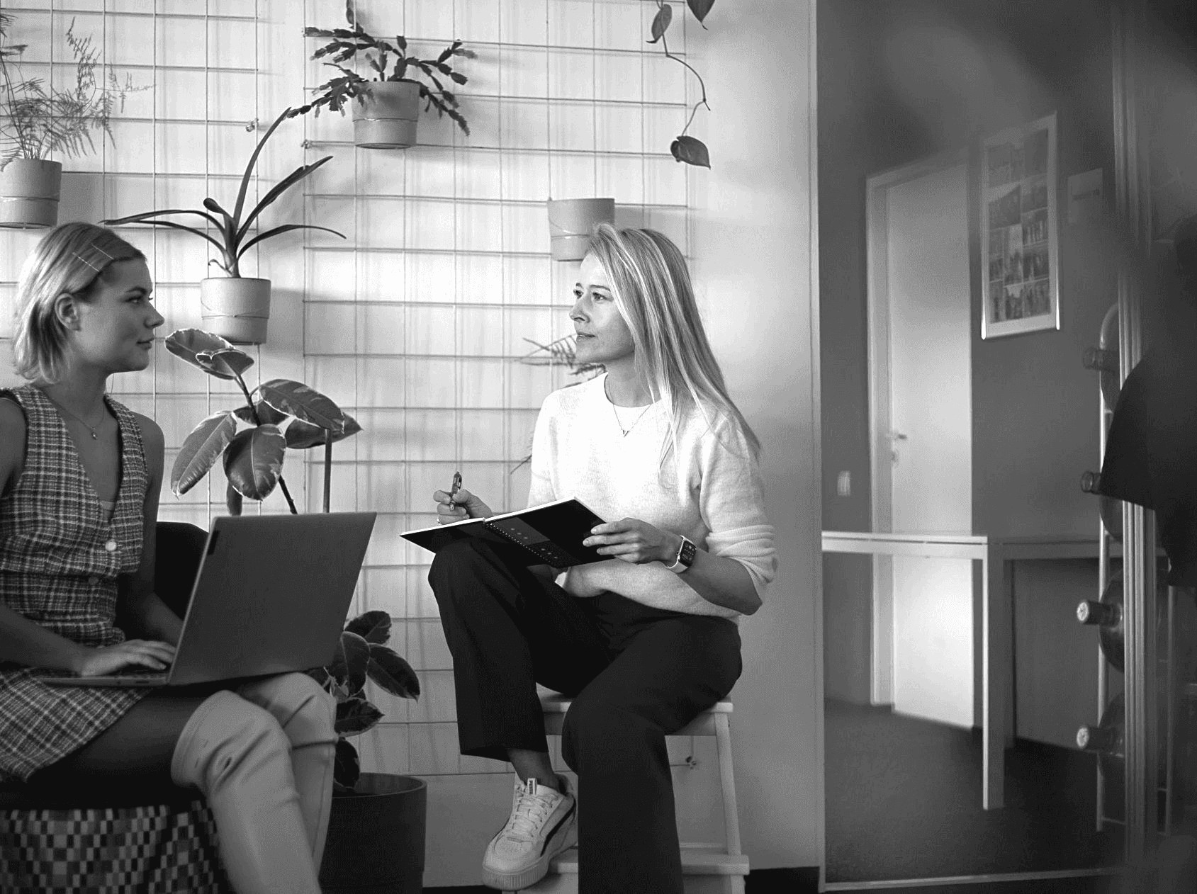 Two people engaged in discussion, one sitting with a laptop and the other holding a notebook, in a modern space.